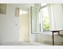 Helles Zimmer mit weißen Wänden, einem offenen Fenster mit Blick nach draußen und einem Heizkörper unter dem Fenster. Im Vordergrund ist ein Bett erkennbar, rechts daneben ein Tisch.