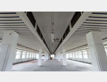 Leerer, langer Raum mit weißen Säulenreihen, einer grauen Betondecke mit Schäden und einer Fensterfront auf beiden Seiten. Der Boden ist unbehandelt, und die Wände tragen minimale Verkleidungen.
