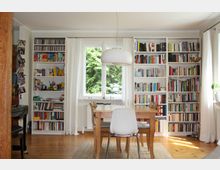 Wohnzimmer mit Holzboden, einem Holztisch mit Stühlen und einer weißen Hängelampe. An der Wand stehen weiße Bücherregale, gefüllt mit bunten Büchern und CDs; in der Mitte des Raums ein Fenster mit Blick ins Grüne, gerahmt von weißen Vorhängen.