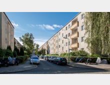 Straßenansicht eines Wohngebiets mit mehrstöckigen, hellen Wohngebäuden im typischen Stil der Nachkriegszeit. Die Gebäude verfügen über Balkone, und die Straße ist von parkenden Autos und Motorrädern sowie grüner Bepflanzung gesäumt.