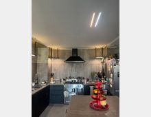 Moderne Küche mit dunklen Schränken, einer Edelstahlkochstelle mit Herd und Dunstabzugshaube in der Mitte. Der Raum hat Betonwände, eine freistehende Kücheninsel mit einem roten Obstständer, und sichtbare Utensilien sowie einen seitlich stehenden Kühlschrank mit Magneten und Dekorationen.