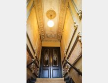 Treppenaufgang in einem historischen Gebäude mit verzierten Wänden und einem reich dekorierten Deckenfries. Oben führt eine doppelflügelige Holztür mit Glaseinsätzen in einen weiteren Raum; eine Pendelleuchte mit Kugel hängt zentral von der Decke.