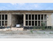 Verlassenes, einstöckiges Gebäude mit beschädigter Fassade und großen, vergitterten Fensteröffnungen. Im Inneren sind Schutt und alte Gegenstände sichtbar, außen wachsen vertrocknete Pflanzen entlang der Wand.