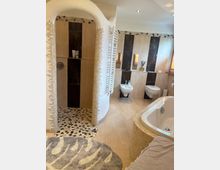 Modernes Badezimmer mit einer offenen Dusche mit Mosaikboden, einer freistehenden Badewanne und beigefarbenen Fliesen mit dunklen Akzenten. Im Hintergrund sind ein Bidet, eine Toilette und kleine Fenster mit Holzjalousien zu sehen.