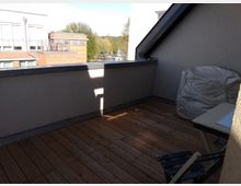 Kleine Dachterrasse mit Holzboden, einer niedrigen Brüstung und einem schrägen Dach. Im Vordergrund stehen ein zusammenklappbarer Stuhl, ein Tisch sowie ein Sitzsack, im Hintergrund sind benachbarte Gebäude und Bäume sichtbar.