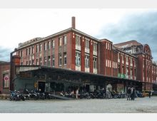 Backstein-Gebäude im Industriedesign mit großen Fenstern und hoch aufragendem Schornstein. Der Vorplatz ist gepflastert, mit geparkten Motorrädern und einer kleinen Menschenansammlung unter einem überdachten Bereich.