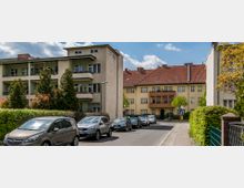 Straße in einem Wohngebiet mit geparkten Autos entlang des Bordsteins. Im Hintergrund stehen Mehrfamilienhäuser mit hellen Fassaden, roten Ziegeldächern und vereinzelten Balkonen, umgeben von Bäumen und Hecken.