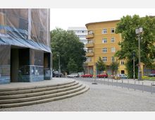 Ein städtischer Platz mit gepflastertem Boden, einer breiten Treppe und einem Gebäude, das teilweise von Bauplanen verdeckt ist, rechts daneben ein gelbes Wohngebäude mit Balkonen und großen Fenstern. Im Hintergrund sind Bäume und parkende Autos zu sehen.