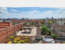 Dachterrasse mit Holzboden, Pflanzkästen und Sitzbereich, umgeben von roten Ziegeldächern und einem Ausblick auf die Stadt mit modernen und historischen Gebäuden im Hintergrund. Vegetation und Begrünung auf Teilen der Dachfläche sichtbar.