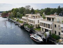 Moderne Reihenhäuser direkt an einem Flussufer mit großzügigen Terrassen und Balkonen, teilweise auf Stelzen über dem Wasser gebaut. Im Vordergrund liegt ein Motorboot im Anlegebereich, während im Hintergrund eine grüne Uferlandschaft und eine rote Brücke zu sehen sind.