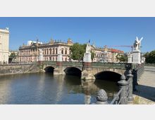 Brücke aus Stein mit grünen Geländern und Statuen auf steinernen Sockeln über einem Fluss, im Hintergrund ein barockes Gebäude in Rosa mit kunstvollen Verzierungen und einer Reihe von Figuren auf dem Dach. Klarer Himmel und Baumreihe an der Seite, rechts im Bild ein gepflasterter Weg.