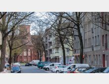 Straßenszene in einer Stadt mit parkenden Autos entlang des Straßenrandes, flankiert von mehrstöckigen Wohngebäuden. Im Hintergrund ist eine Backsteinkirche mit großen, gotischen Fenstern sichtbar, umgeben von Bäumen mit vereinzeltem Laub.