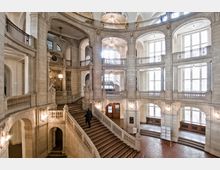 Großes, historisches Treppenhaus mit symmetrischen Bögen, Balustraden und Statuen, gestaltet aus hellgrauem Stein. Hohe Fenster lassen viel Tageslicht herein, und Kronleuchter betonen die klassizistischen Details der Innenarchitektur.