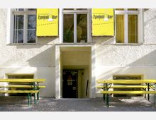 Außenansicht eines Gebäudes mit gelben Schildern der "Zyankali-Bar" über Fenstern; darunter ein Eingang in einen Keller mit einer dunklen Tür, die Warnschilder trägt. Vor dem Gebäude stehen gelbe Bänke mit grünen Metallgestellen auf einem gepflasterten Boden.