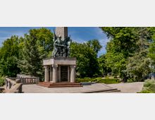 Denkmal mit einer Bronze-Skulptur von Soldaten auf einem rechteckigen Sockel vor einem Obelisken, umgeben von Bäumen und gepflegten Gartenanlagen mit Kieswegen. Im Hintergrund sind weitere Parkstrukturen und ein Pavillon zu sehen.