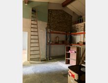 Interior of a warehouse or storage room with a concrete floor, exposed brick and plaster walls, and a high sloped ceiling. A tall wooden ladder leans against the wall beside metal shelving with boxes, with a small mezzanine railing and scattered furniture pieces in the foreground.