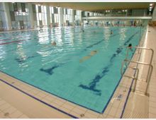 Innenansicht eines Hallenbads mit einem großen rechteckigen Schwimmbecken, das von hellen Fliesen umgeben ist. Das Becken ist in Bahnen unterteilt, die von blauen Linien markiert sind, und mehrere Personen schwimmen darin; im Hintergrund sind große Fenster und eine Galerieebene sichtbar.