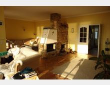 Wohnzimmer mit gelb gestrichenen Wänden, einem zentralen gemauerten Kamin mit weißem Einsatz und einer gläsernen Tür, die zu einem weiteren Raum führt. Der Raum ist mit einem beigen Sofa, einem Sessel und terrakottafarbener Fliesen ausgelegt, ergänzt durch eine helle Teppichfläche und einige Pflanzen.