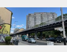 Straßenansicht in einer Stadt mit einer erhöhten Stahlbahntrasse, einem großen Wohngebäude im Hintergrund und Verkehrszeichen für die Kreuzung Gitschiner Straße und Segitzdamm. Im Vordergrund sind ein bemaltes Haus, parkende und fahrende Autos sowie Gehwege mit Pflanzenelementen zu sehen.