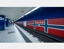 U-Bahn-Station "Osloer Straße" in Berlin mit einem Bahnsteig aus hellem Bodenbelag, roten Wandpaneelen mit blauen und weißen Kreuzmustern, die an die norwegische Flagge erinnern. Beleuchtung und Informationsanzeigen befinden sich an der Decke, während Personen in Richtung des Ausgangs gehen.