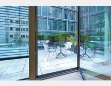 Blick durch eine Glasfassade auf eine moderne Terrasse mit weißen und schwarzen Stühlen sowie kleinen Tischen, umgeben von Pflanzen. Im Hintergrund befinden sich hohe, verglaste Bürogebäude.