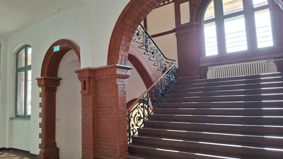 Innenansicht eines historischen Treppenhauses mit breiter Holztreppe, kunstvollem schmiedeeisernem Geländer und roten Backsteinbögen. Große Rundbogenfenster lassen viel Licht einfallen, darunter steht ein Heizkörper an der Backsteinwand.