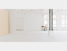 Weißer, minimalistisch gestalteter Innenraum mit großen Fenstern, die viel Tageslicht hereinlassen. Im Raum befinden sich eine Metallstütze, eine kleine Tisch-Stuhl-Kombination mit einem Laptop und eine locker gespannte Lichterkette an der Decke.