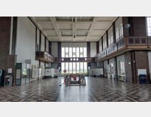 Großer, lichtdurchfluteter Innenraum mit hohen Decken im Stil der frühen Moderne, geprägt von Backsteinwänden, großen Fenstern und einer umlaufenden Galerie. Der Boden ist mit einem geometrischen Muster gefliest, und es sind Vitrinen, Ausstellungstafeln und Sitzgelegenheiten verteilt.