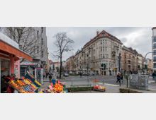 Straßenkreuzung in einer städtischen Wohngegend mit mehrstöckigen Altbauten im Gründerzeitstil. Im Vordergrund befindet sich ein Marktstand mit Obst und Gemüse, im Hintergrund sind Geschäfte, Fußgänger und parkende Autos zu sehen.