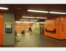 U-Bahn-Station mit orangefarbenen Wänden und Decken, gefliestem Boden und Neonbeleuchtung. Im Hintergrund führen Rolltreppen und eine Treppe nach unten, die Wände sind teils mit Graffiti bedeckt.
