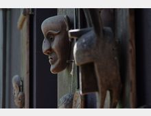 Nahaufnahme von mehreren hölzernen Masken, die an einer Wand aus Holz und dunklem Material befestigt sind. Die Masken zeigen unterschiedliche Gesichtsformen und wirken handgeschnitzt.