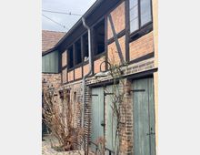 Fachwerkgebäude mit einer Fassade aus Ziegelstein und dunkelbraunen Holzbalken, daneben grüne Holztüren im Erdgeschoss. Im Vordergrund sind kahle Sträucher und ein gepflasterter Hof zu sehen.