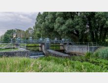 Eine kleine Schleusenanlage aus Beton mit metallenen Geländern und Toren, umgeben von dichten Bäumen und üppigem Grün. Ein Wasserlauf fließt durch die Schleuse, während das Gelände ländlich und naturbelassen wirkt.