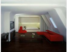 Moderne Dachgeschosswohnung mit minimalistischem Design, bestehend aus einem offenen Wohnbereich mit rotem Sofa und Sessel, dunklem Holzboden und einem eingebauten Bett im Hintergrund. Ein Schreibtisch, ein Fernseher und schräge Decken mit Dachfenstern bestimmen die Raumgestaltung.