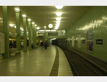 U-Bahn-Station mit grünen Fliesenwänden und Metallstützen, beleuchtet durch runde Deckenlampen; eine gebogene Bahnsteigkante führt entlang der Gleise, während im Hintergrund eine Treppe zur oberen Ebene führt. Ein Wanduhr zeigt die Zeit, und an den Wänden sind Werbetafeln sowie Schilder mit der Aufschrift "Alexanderplatz" angebracht.