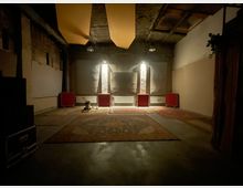 Ein dunkler Raum mit sichtbaren Ziegelwänden und einer hohen Decke. Der Boden ist mit mehreren Teppichen bedeckt, an der Rückwand stehen rote Möbelstücke und Heizkörper, und die Beleuchtung erfolgt durch wenige Wandlampen.