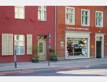 Fassadenansicht von zwei miteinander verbundenen Gebäuden aus rotem Backstein mit großen Fenstern; links eine dekorative Holztür und Fenster mit beigefarbenen Fensterläden, rechts ein Schaufenster mit der Aufschrift „Coiffeur Schnittpunkt“. Vor den Gebäuden stehen Blumenkästen und Fahrradbügel auf dem Gehweg.