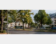 Straßenszene in einem Wohngebiet mit mehrstöckigen Wohnhäusern im Hintergrund, umgeben von großen Bäumen. Am Straßenrand stehen geparkte Autos, ein Bus hält an einer Haltestelle, und ein Straßenschild kennzeichnet den "Göllweg".