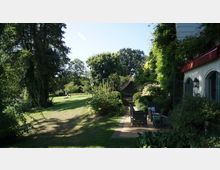 Ein gepflegter Garten mit Rasenfläche, hohen Bäumen und dichter Vegetation, die eine Terrasse mit Gartenmöbeln an einem Haus mit begrüntem Fassadenbereich umgibt. Rechts ist ein Teil der Hauswand mit großen Fenstern und einer roten Markise sichtbar.