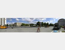 Weitläufiger Stadtplatz mit gepflastertem Boden, umgeben von mehreren Gebäuden, darunter ein moderner Wohnblock links und ein historisches Kuppelgebäude rechts. In der Mitte eine Kreuzung mit Bäumen, parkenden Autos und einem kleinen Kiosk im Vordergrund.