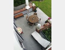 Ansicht einer Terrasse mit grauem Bodenbelag, einem runden Holztisch und mehreren Stühlen in der Mitte. Im Hintergrund befinden sich eine Ziegelmauer mit Pflanzkübeln sowie ein angrenzender Garten mit Rasenfläche und Hecken. Links sind eine Holzbank, ein kleiner Beistelltisch und eine gepolsterte Liege zu sehen.