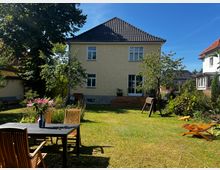 Ein Garten mit Rasen und vereinzelten Bäumen vor einem zweistöckigen, hellgelben Haus mit Satteldach und weißen Fenstern. Im Vordergrund stehen ein Holztisch mit Stühlen und eine Vase mit rosa Blumen, umgeben von Gartenmöbeln und Spielgeräten.