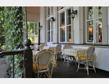 Eine Außenterrasse mit weiß gerahmten Fenstern und mehreren Sitzgruppen aus geflochtenen Stühlen und Tischen, die mit gelb-weiß karierten Tischdecken bedeckt sind. Eine Wand ist mit grünen Kletterpflanzen bewachsen, und die Umgebung ist von Bäumen umgeben.