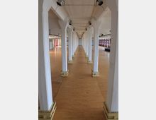 Langer, rechteckiger Innenraum mit hellen Holzböden und weißen Säulenreihen, die von der Decke zum Boden verlaufen. An der Decke sind Scheinwerfer angebracht, und große Fenster auf einer Seite lassen Tageslicht herein.