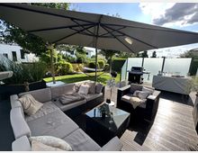 Moderne Terrasse mit Sitzecke aus Polyrattan-Möbeln und grauen Polstern, überdacht von einem großen Sonnenschirm. Im Hintergrund befinden sich ein Garten mit Rasen und Bäumen, eine Hängematte sowie eine Grillstation vor einer teilmatten Sichtschutzwand.
