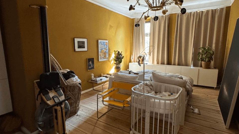 Cozy bedroom with mustard-yellow walls, wide wooden plank flooring, and a large mid-century style chandelier on the ceiling. A bed sits in front of a curtained window beside a long white cabinet with potted plants, with a crib and chairs in the foreground.