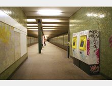 Ein unterirdischer U-Bahnsteig mit langen Fliesenwänden in Grün, von Neonlicht beleuchtet. Im Vordergrund stehen Fahrkartenautomaten, die mit Graffiti bedeckt sind, während im Hintergrund Sitzbänke und eine digitale Anzeigetafel sichtbar sind.