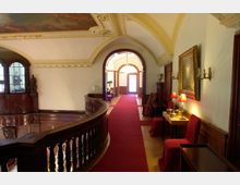 Obergeschoss eines historischen Gebäudes mit gekrümmtem Holzgeländer, rotem Teppich und gewölbter Decke mit Stuckverzierungen. An den Wänden befinden sich Gemälde und Beleuchtung, im Hintergrund führt ein offener Durchgang zu weiterem Tageslicht.