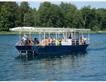 Ein kleines Ausflugsboot mit offenem Dach und Passagieren fährt auf einem ruhigen See, umgeben von Wald und grüner Vegetation.