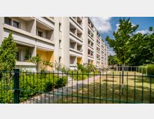 Mehrstöckiger Wohnblock mit Balkonen in einer hellen, teils gelben Fassade, umgeben von gepflegten Grünflächen. Im Vordergrund ein Metallzaun, dahinter ein Gartenbereich mit Bäumen, Rasen und einem gepflasterten Weg.